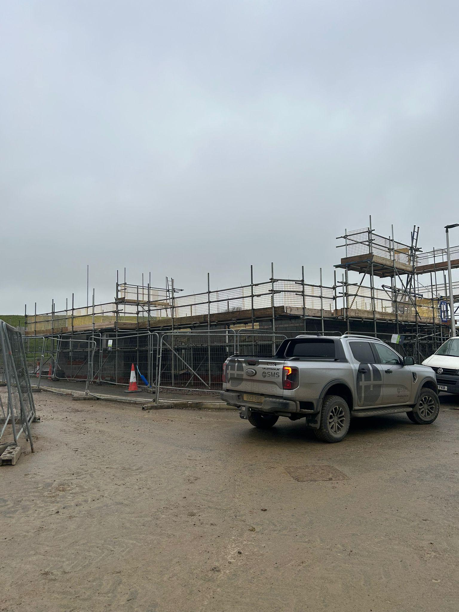 A construction site with domestic scaffolding, a parked pickup lorry, and a cloudy sky overhead.