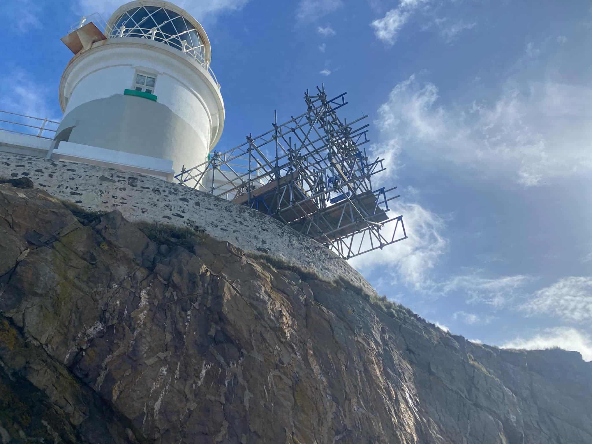 White lighthouse with scaffolding stands on a rocky cliff under a partly cloudy blue sky.