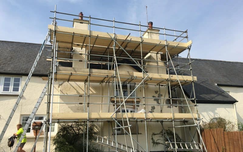 A house with specialist scaffolding and ladders set up for refurbishment; a worker stands nearby.